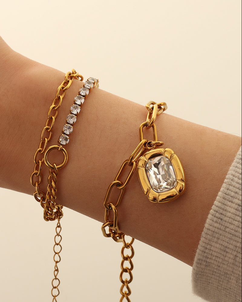 Pulsera dorada de eslabones alargados y piedra transparente rectangular, puesta en la muñeca junto a otras pulseras.