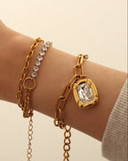 Pulsera dorada de eslabones alargados y piedra transparente rectangular, puesta en la muñeca junto a otras pulseras.