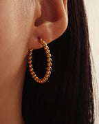 Pendiente dorado de aro formado por pequeñas esferas, puesto en la oreja junto al cabello oscuro.