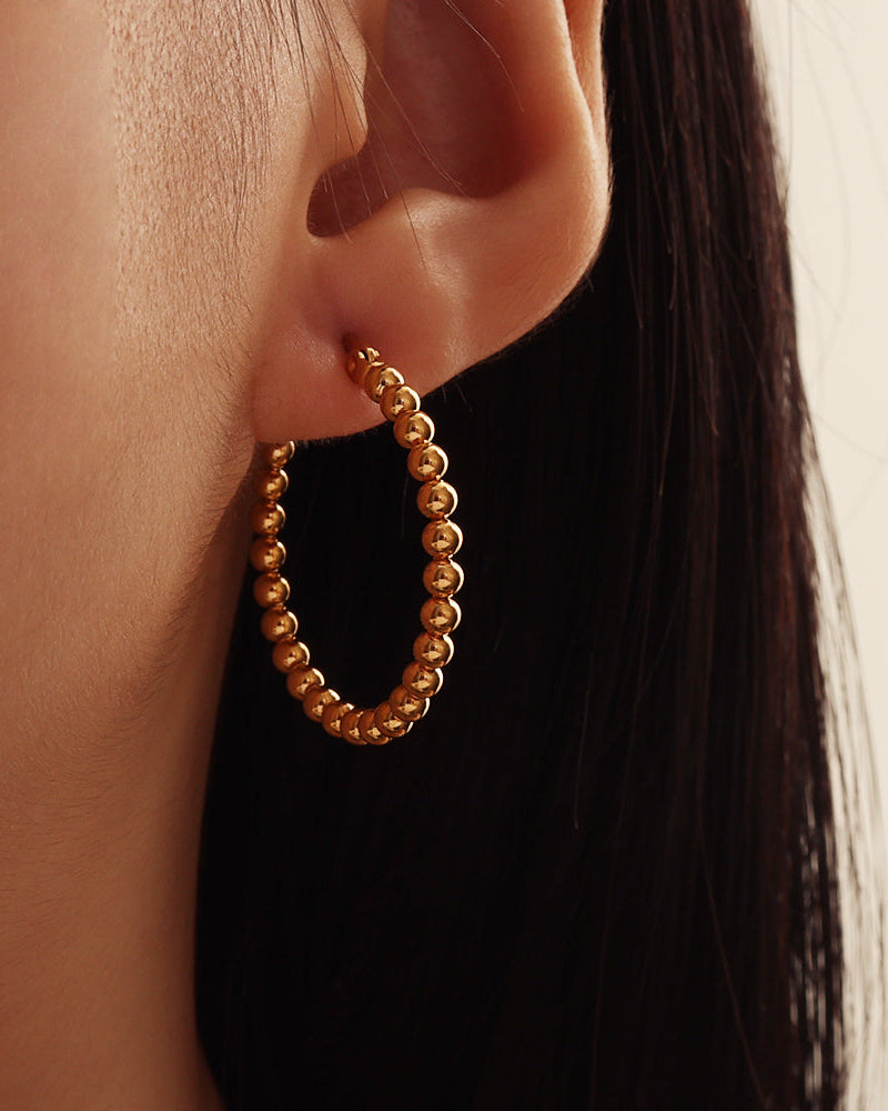 Pendiente dorado de aro formado por pequeñas esferas, puesto en la oreja junto al cabello oscuro.