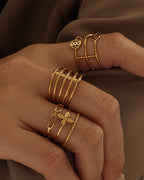 Dos manos con varios anillos dorados de bandas finas, pequeños círculos, corazones y diseños calados, sobre tela oscura.