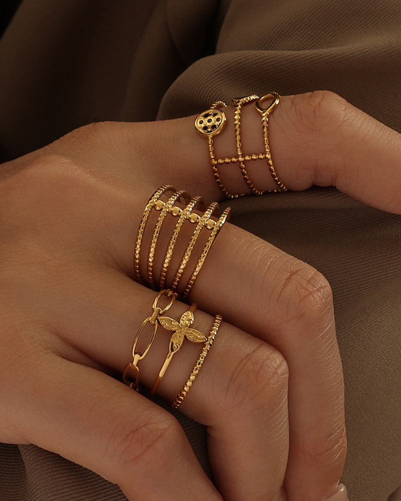 Dos manos con varios anillos dorados de bandas finas, pequeños círculos, corazones y diseños calados, sobre tela oscura.