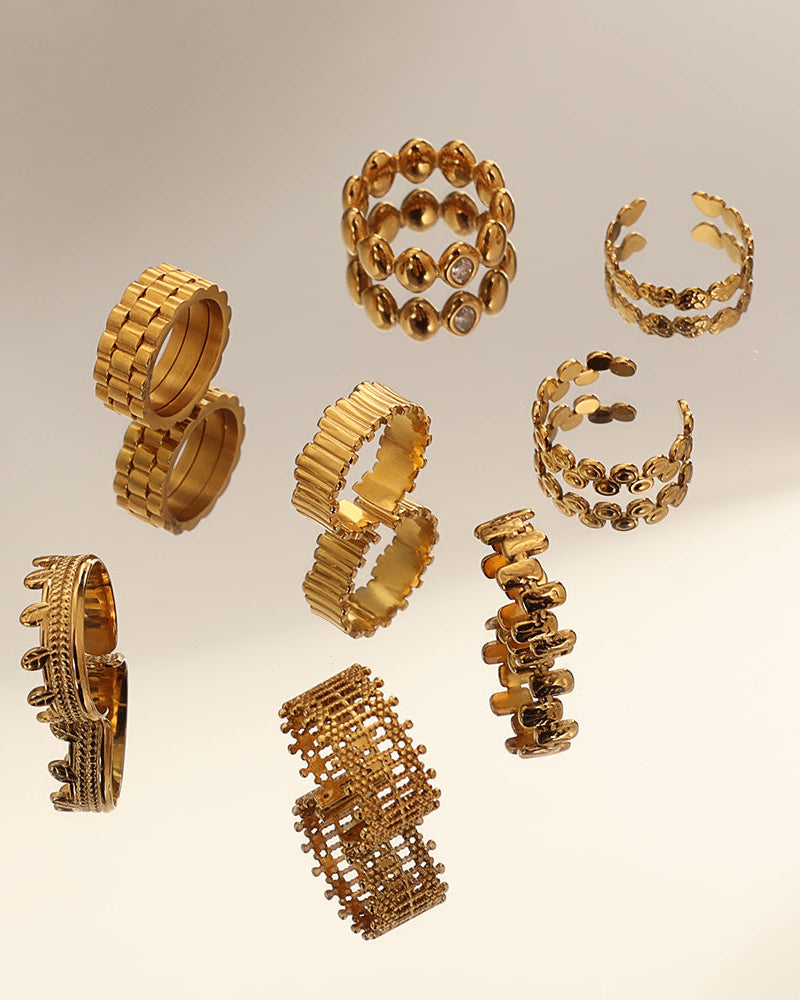 Varios anillos dorados de bandas anchas, cadenas y diseños calados, sobre superficie reflectante.
