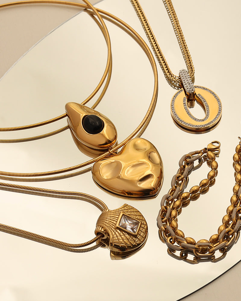 Varias joyas doradas sobre superficie reflectante: colgantes de corazón, gota y círculo, junto a una pulsera de eslabones.