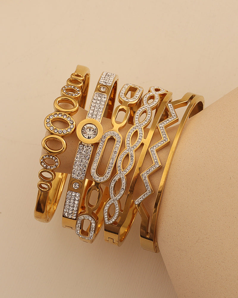 Varias pulseras rígidas doradas con círculos, óvalos y zigzag de pequeñas piedras claras, sobre un soporte beige.