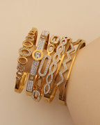 Varias pulseras rígidas doradas con círculos, óvalos y zigzag de pequeñas piedras claras, sobre un soporte beige.