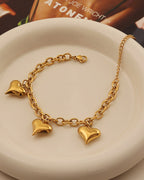 Pulsera dorada de cadena con tres corazones lisos colgantes, sobre un plato blanco.