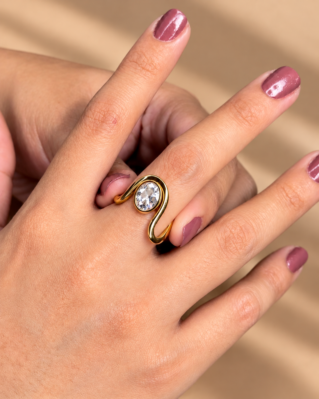 Anillo abierto dorado de banda sinuosa y piedra transparente ovalada, puesto en una mano con uñas moradas.