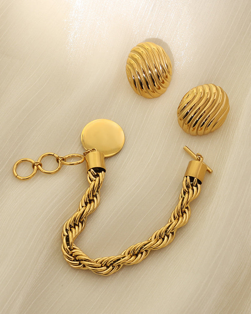 Pulsera dorada trenzada con disco liso colgante y cierre de barra, junto a pendientes redondos estriados.