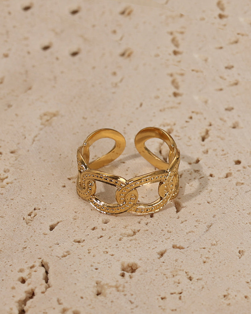 Anillo abierto dorado de grandes eslabones ovalados con pequeñas piedras claras, sobre piedra beige.