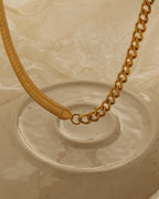 Collar dorado con una mitad de cadena plana y otra de eslabones, dispuesto sobre un plato de cerámica claro.