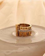 Anillo ancho dorado con números romanos oscuros y franja de pequeñas piedras claras, sobre plato de cerámica.