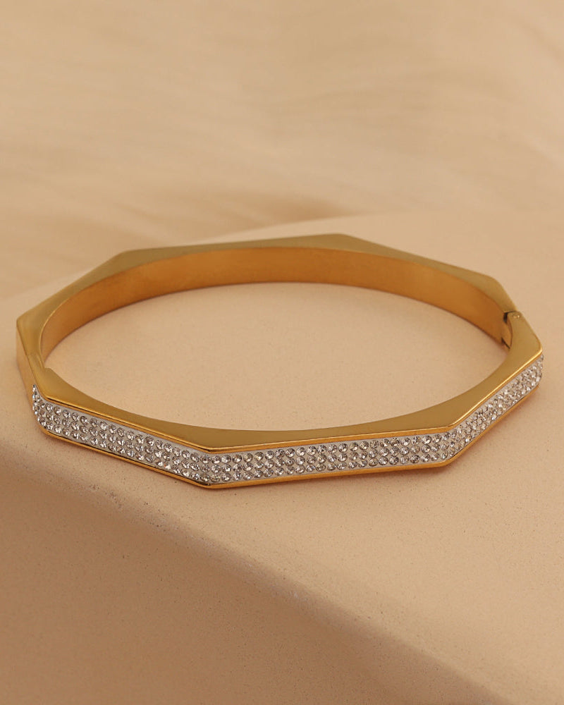 Pulsera rígida dorada de forma poligonal con varias hileras de pequeñas piedras claras en el frente, sobre tela beige.