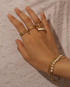 Mano con varios anillos dorados y pulseras finas, una de perlas blancas, sobre mármol rosado.