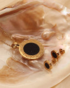 Collar y pendientes dorados de motivos redondos negros y bordes grabados, sobre una concha de tonos crema.