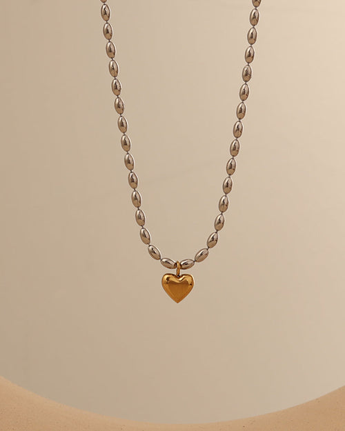 Collar de cuentas ovaladas de tono plateado con pequeño corazón dorado colgante, sobre fondo beige.