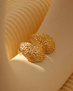 Par de pendientes dorados redondeados y abombados con textura granulada, sobre soporte beige iluminado por el sol.