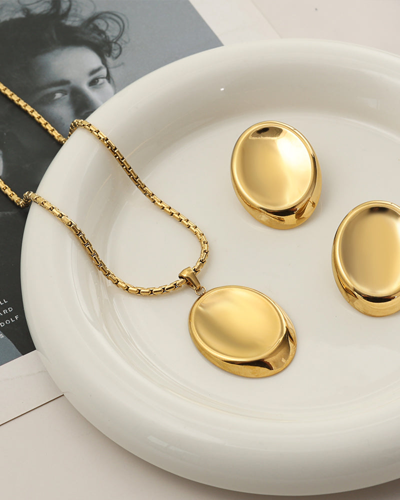 Collar dorado de colgante ovalado liso con pendientes a juego sobre plato blanco y fotografía.