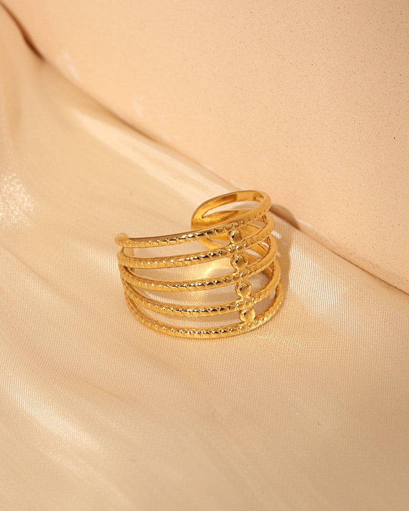 Anillo abierto dorado de varias bandas paralelas con pequeñas piedras claras, sobre tela beige.