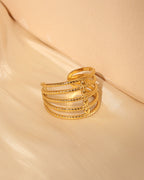 Anillo abierto dorado de varias bandas paralelas con pequeñas piedras claras, sobre tela beige.
