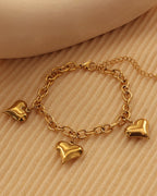 Pulsera dorada de eslabones ovalados con tres corazones colgantes y cadena de extensión, sobre tela beige.