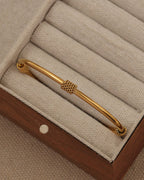 Pulsera rígida dorada con adorno central de textura entrelazada, en un joyero de tela beige.