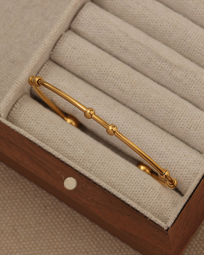 Pulsera rígida dorada de aro fino con pequeñas esferas, en un compartimento de joyero de tela beige.