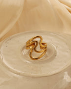 Anillo dorado ancho de líneas curvas onduladas y espacios abiertos, sobre un plato de cerámica claro.