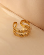 Anillo abierto ancho dorado con bandas onduladas entrecruzadas y huecos alargados, sobre tela beige.