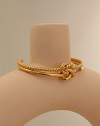 Pulsera rígida dorada de dos bandas con textura de cuerda y nudo central, sobre soporte beige.