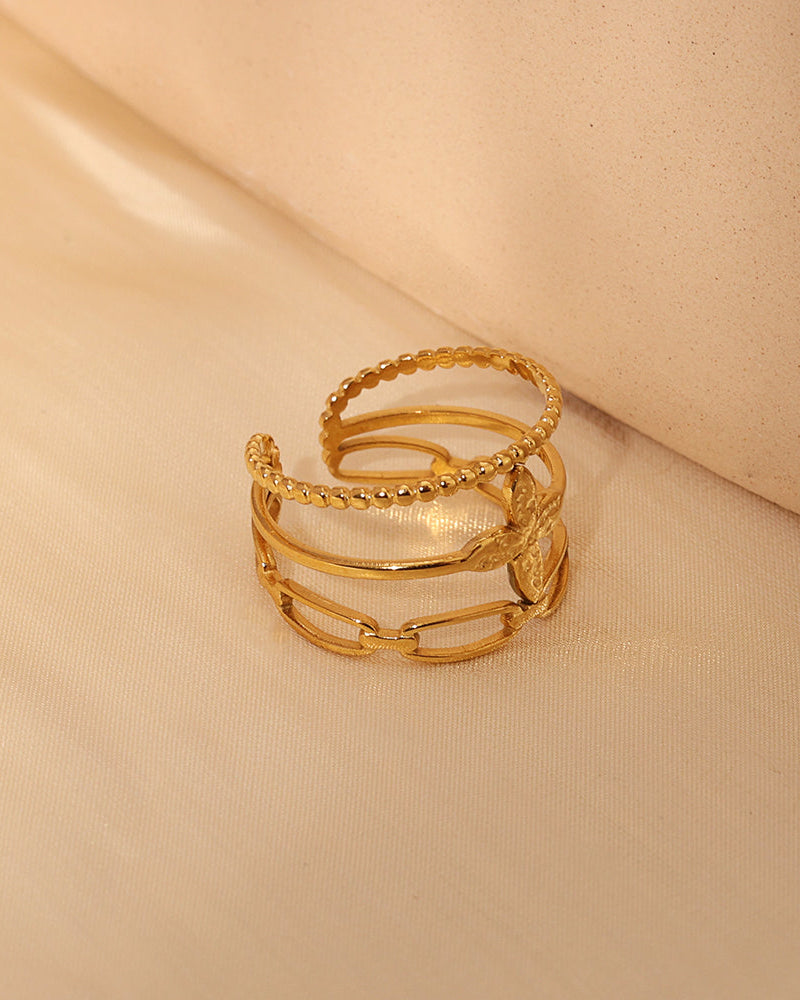 Anillo abierto dorado de varias bandas con texturas, motivos circulares y formas geométricas, sobre tela beige.