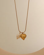 Collar fino dorado con colgante de corazón con textura y perla blanca, sobre fondo claro.