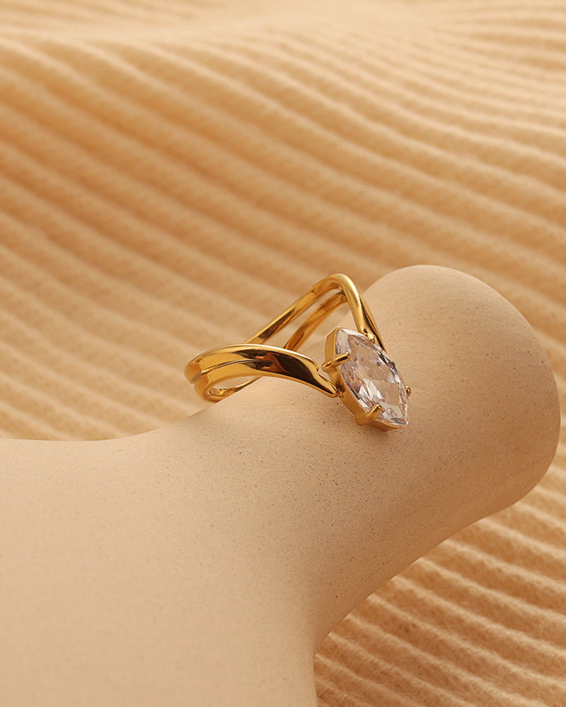 Anillo abierto dorado de bandas curvas con piedra transparente de forma alargada, sobre soporte beige.