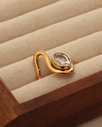 Anillo abierto dorado de banda curva con piedra transparente ovalada, en un joyero de tela beige.