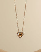 Collar dorado con colgante de corazón de centro dorado y contorno de pequeñas piedras claras, sobre fondo beige.