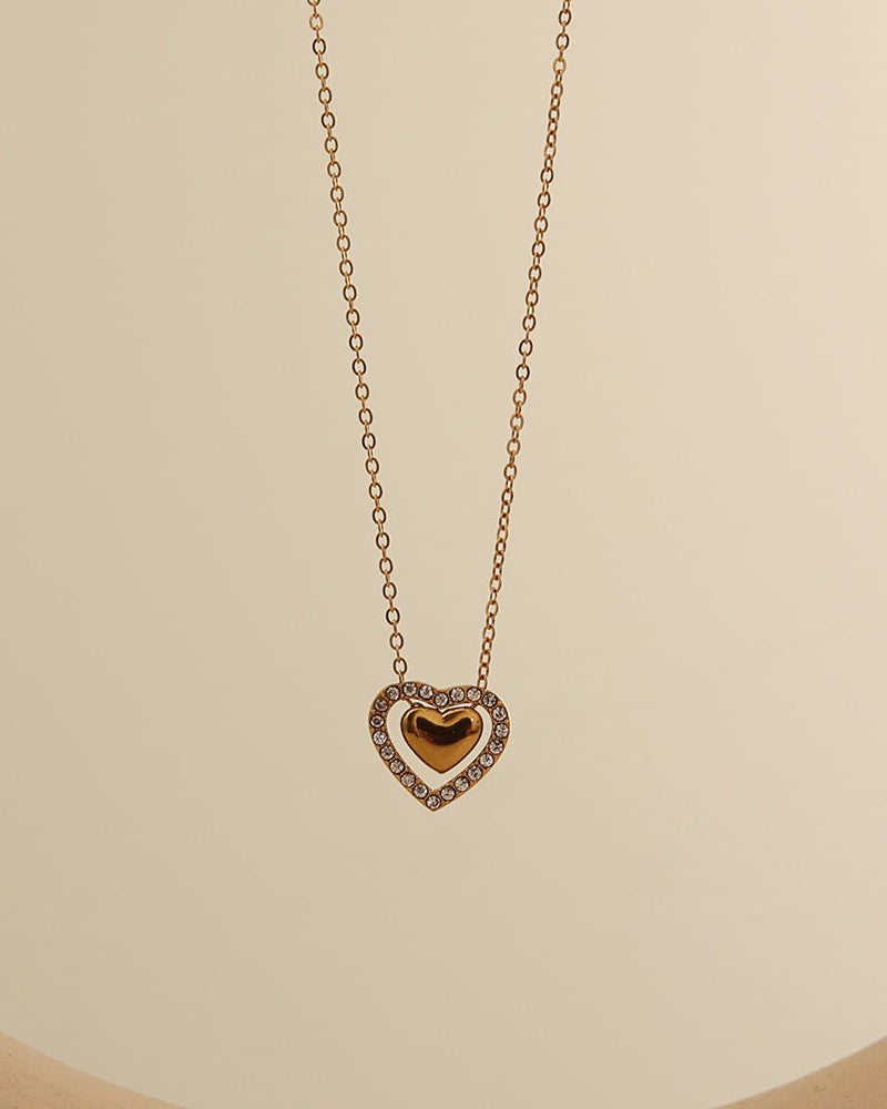 Collar dorado con colgante de corazón de centro dorado y contorno de pequeñas piedras claras, sobre fondo beige.