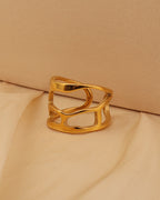 Anillo ancho dorado calado con formas curvas entrecruzadas, sobre tela beige.