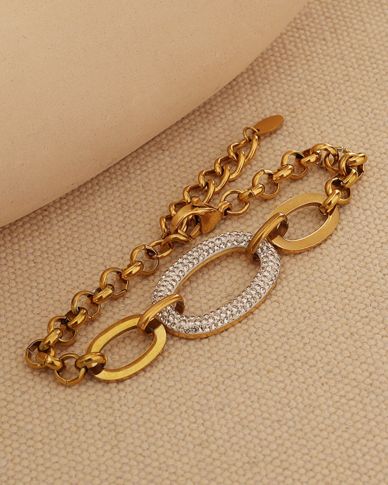 Pulsera dorada de cadena con grandes eslabones ovalados centrales, uno cubierto de pequeñas piedras claras, sobre tela beige.