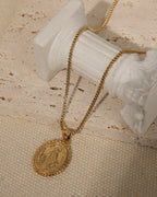 Collar dorado con medallón ovalado de borde ornamentado, dispuesto junto a una columna blanca sobre piedra y tela beige.