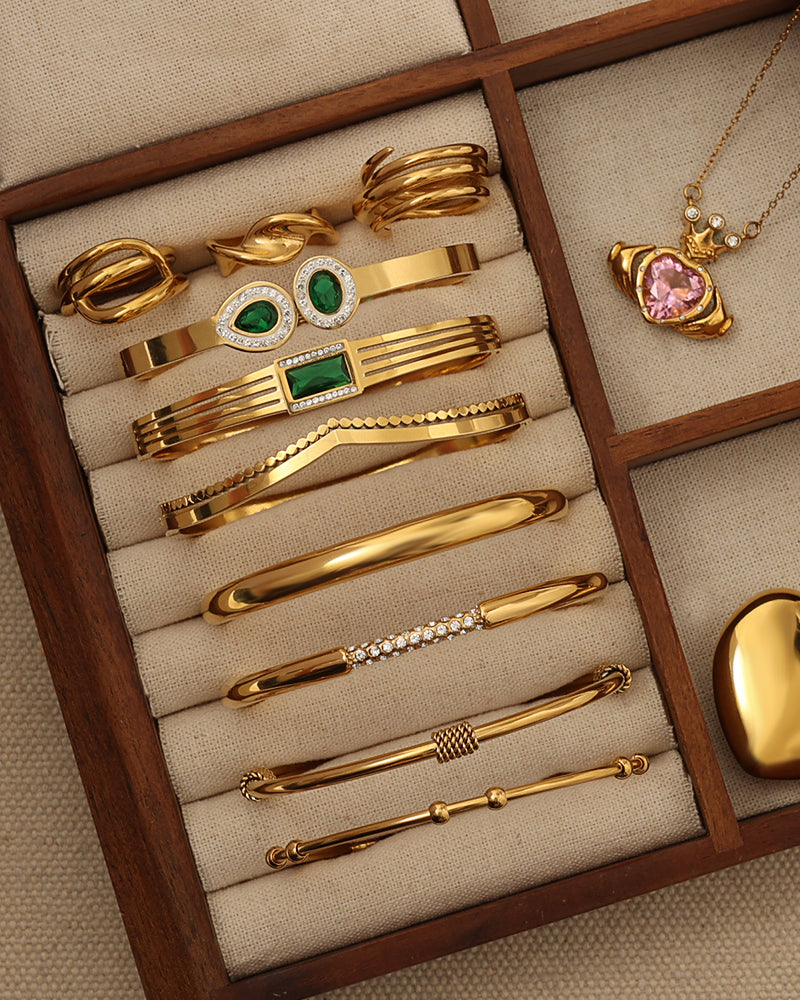 Joyero con varias pulseras rígidas doradas, algunas con piedras verdes o detalles entrelazados, y un collar rosa.