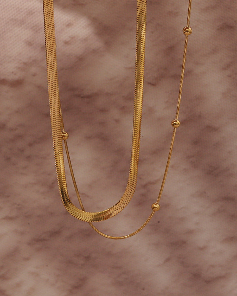 Collar doble dorado con cadena plana y cadena fina con pequeñas esferas, sobre fondo de mármol rosado.