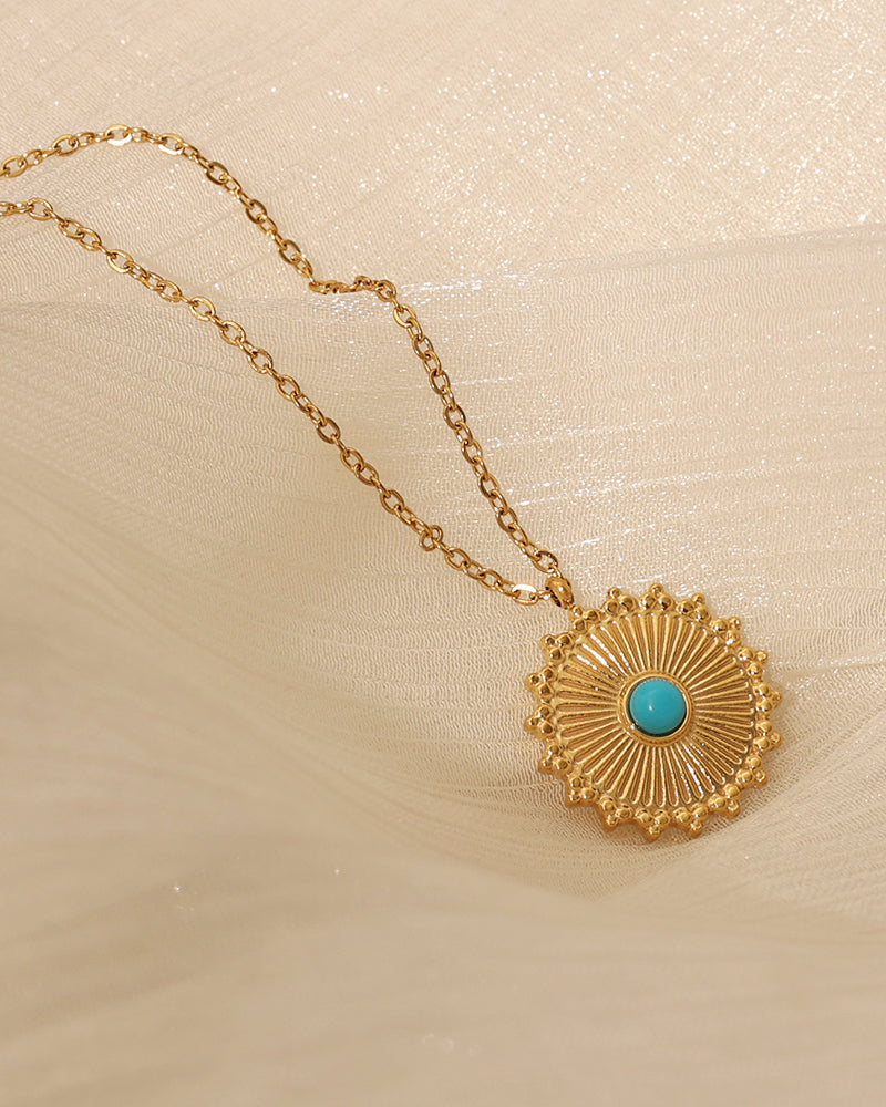 Colgante circular dorado con rayos y pequeño centro azul turquesa, sobre tela clara translúcida.