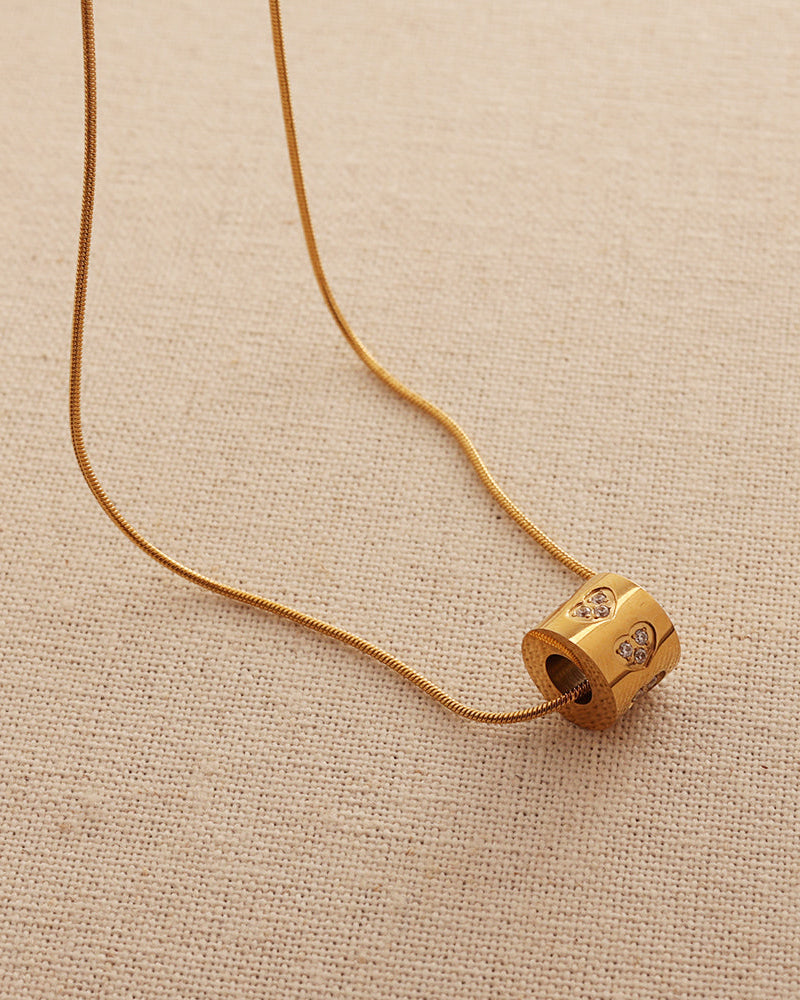 Collar fino dorado con colgante cilíndrico y pequeños motivos de piedras claras, sobre tela beige.