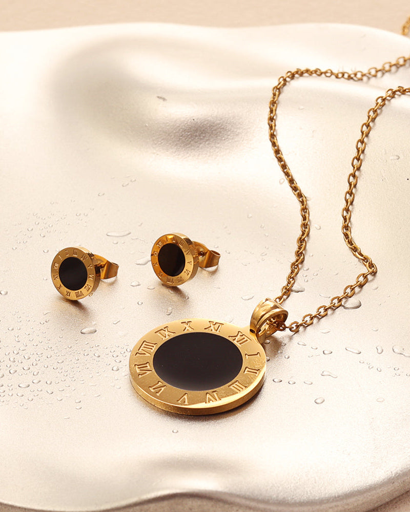 Collar dorado de medallón negro con números romanos y pendientes a juego, sobre superficie satinada clara.