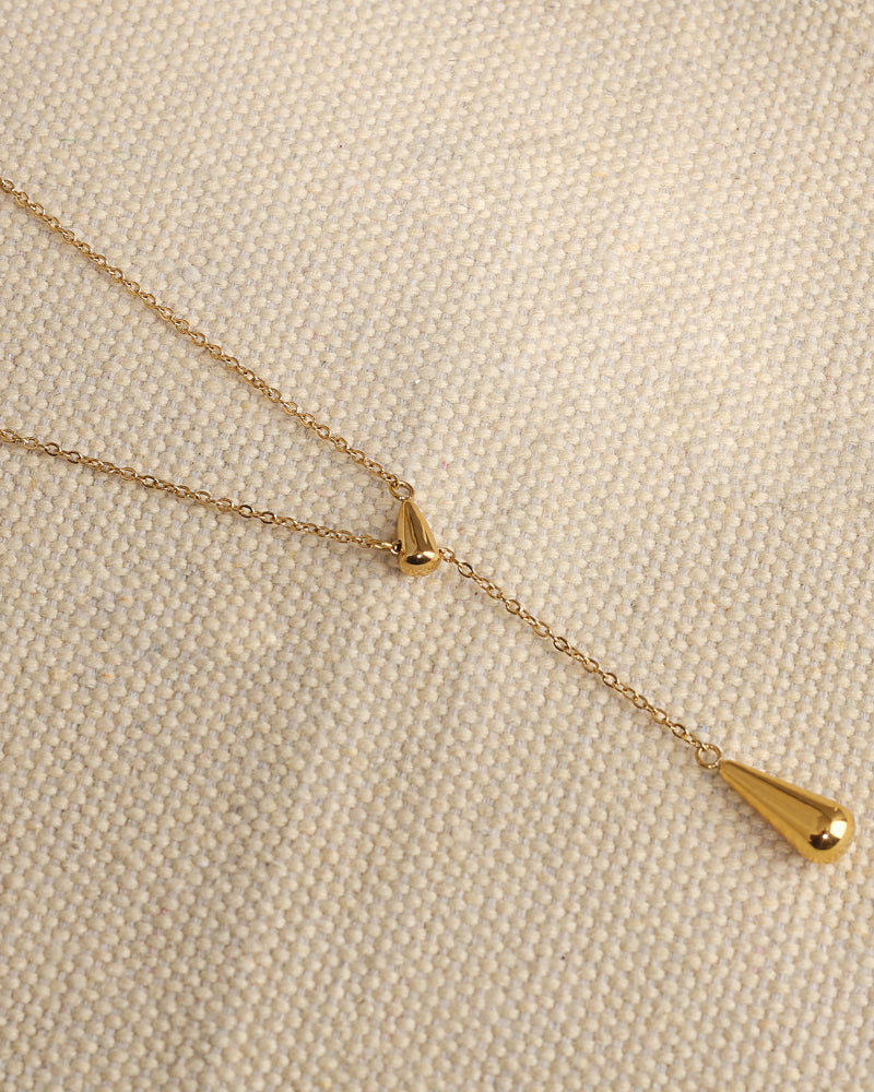 Collar dorado en forma de Y con dos gotas lisas de distinto tamaño sobre tela beige.