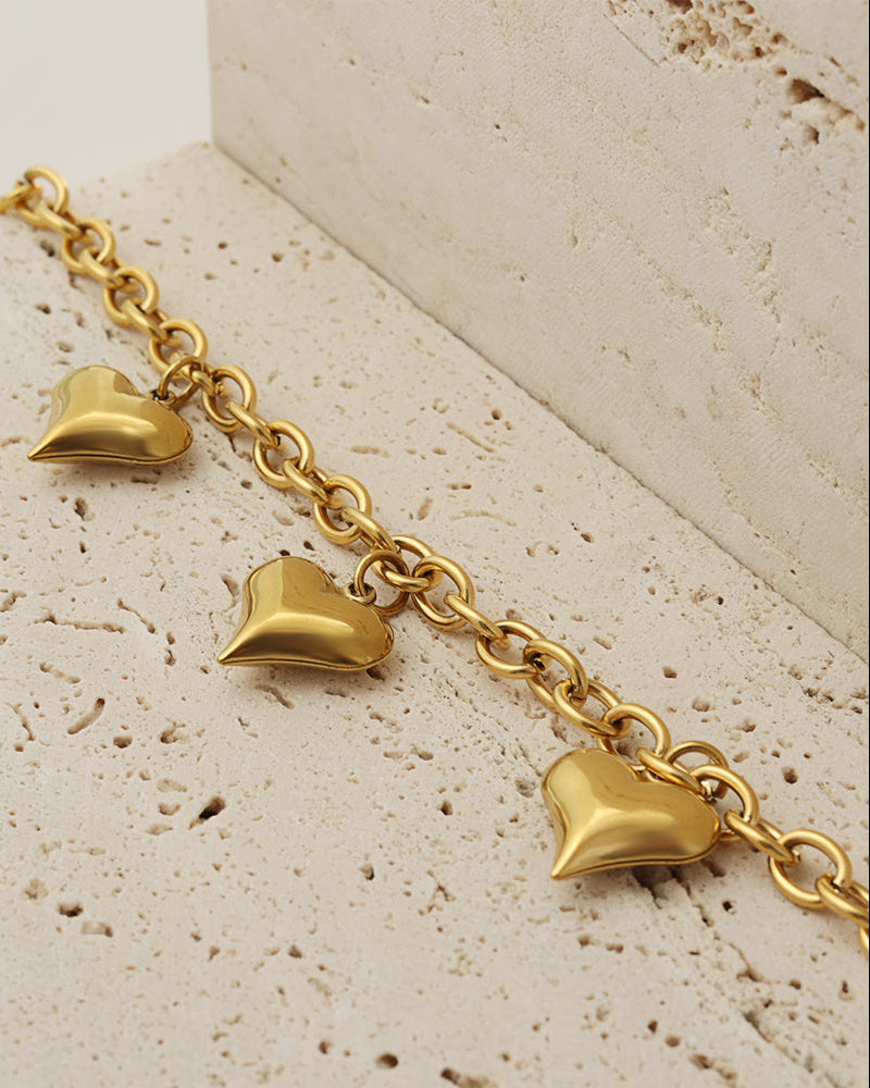 Detalle de pulsera dorada de eslabones ovalados con tres corazones abombados colgantes, sobre piedra clara.