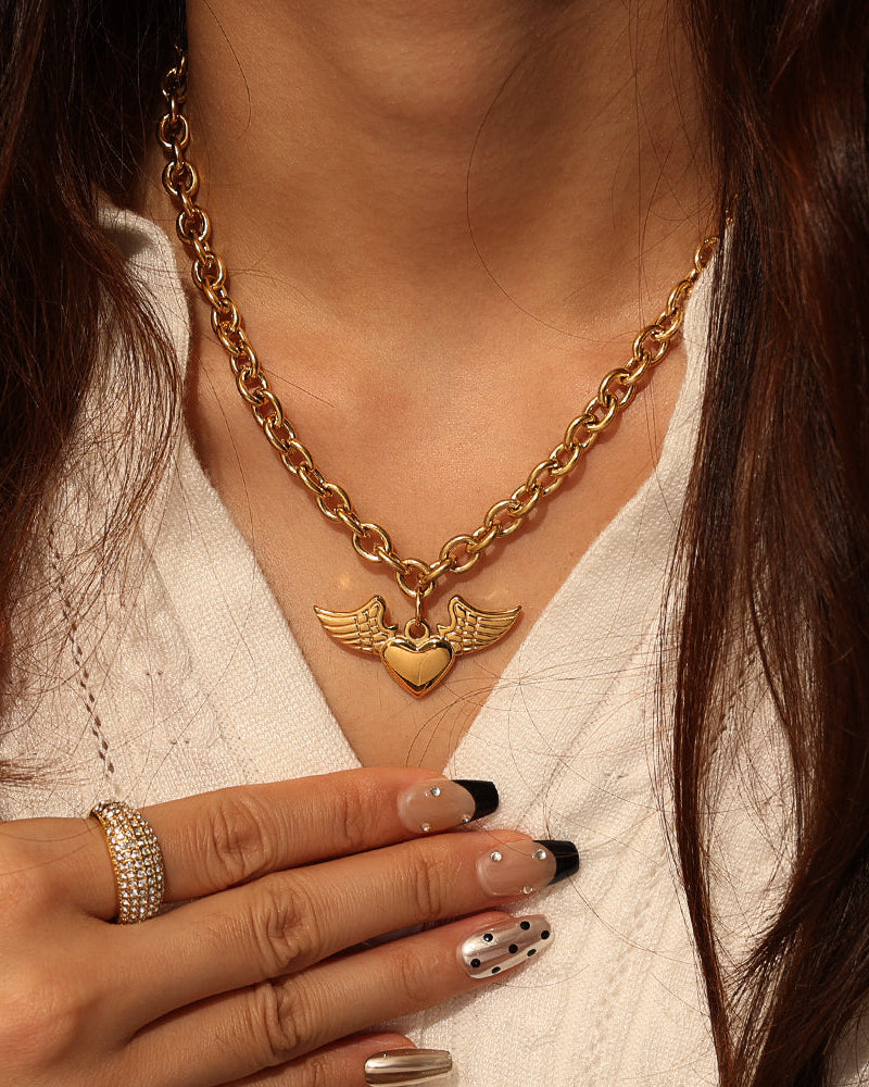 Persona con collar de cadena dorada y colgante de corazón con alas, sobre blusa blanca.
