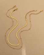 Dos cadenas doradas extendidas sobre tela beige, mostrando sus cierres, una plana y otra fina con pequeñas esferas.
