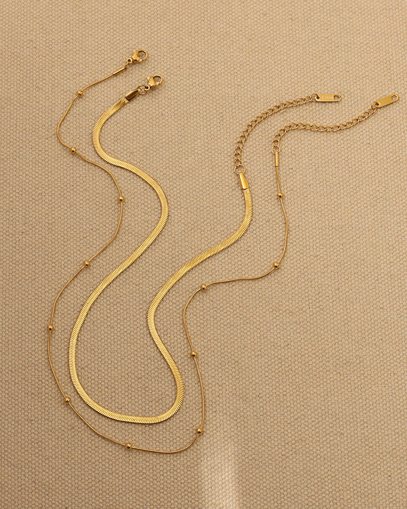 Dos cadenas doradas extendidas sobre tela beige, mostrando sus cierres, una plana y otra fina con pequeñas esferas.