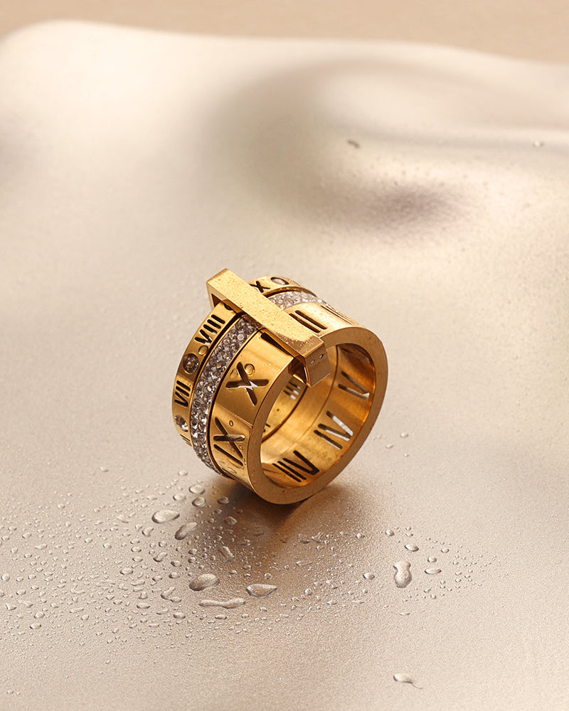 Anillo ancho dorado de varias bandas, números romanos y pequeñas piedras claras, sobre superficie satinada clara.